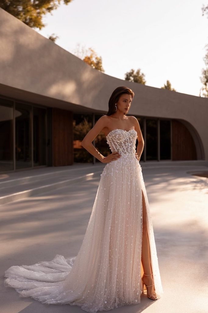 In questa foto, una modella posa indossando un abito della collezione sposa Demetrios 2026 dalla silhouette scivolata in tulle glitter, con spacco laterale, bustier steccato a vista con scollo a cuore e ricami di perline e paillettes