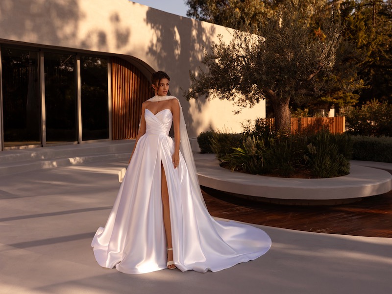 In questa foto, una modella posa indossando un abito della collezione sposa Demetrios 2026 in mikado, con silhouette ad A, spacco frontale e lunga stola al collo