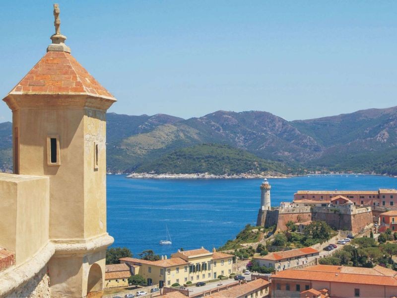In questa foto si vede una suggestiva veduta panoramica dell’Isola d’Elba: in primo piano una torretta in pietra color ocra, sullo sfondo la fortezza medicea di Portoferraio con il suo faro e il mare blu intenso che circonda l’isola. Oltre l’acqua, si stagliano le montagne verdi dell’arcipelago toscano sotto un cielo terso. L’atmosfera è luminosa, mediterranea, ricca di storia e natura.