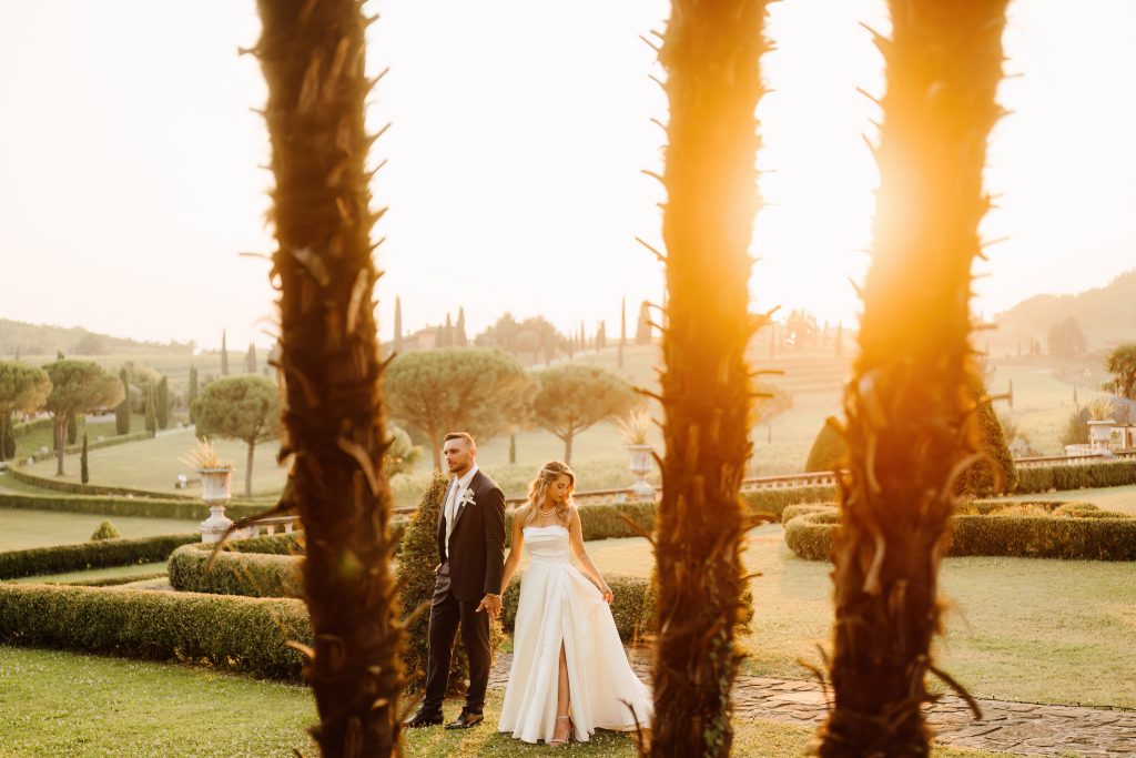 In questa foto, gli sposi sono ritratti al tramonto in un paesaggio da sogno: camminano mano nella mano tra i giardini di una location raffinata, incorniciati da palme e luce dorata. L’atmosfera è romantica e suggestiva. Scatto tratto dal progetto matrimoni 2025 di Ilaria & Andrea.