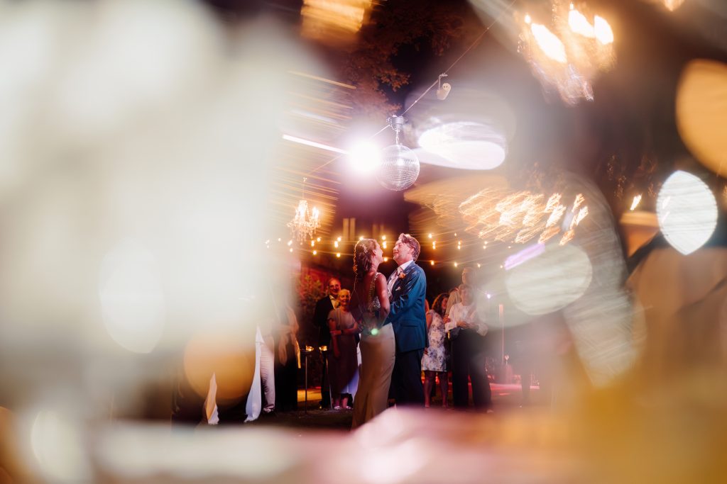 In questa foto, gli sposi danzano sotto le luci calde di una festa all’aperto, tra lampadine sospese e una sfera da discoteca. La scena è avvolta da giochi di luce e riflessi, con gli invitati che osservano sullo sfondo. Un momento magico e vibrante dei matrimoni 2025 di Ilaria & Andrea, dove la luce racconta emozioni.