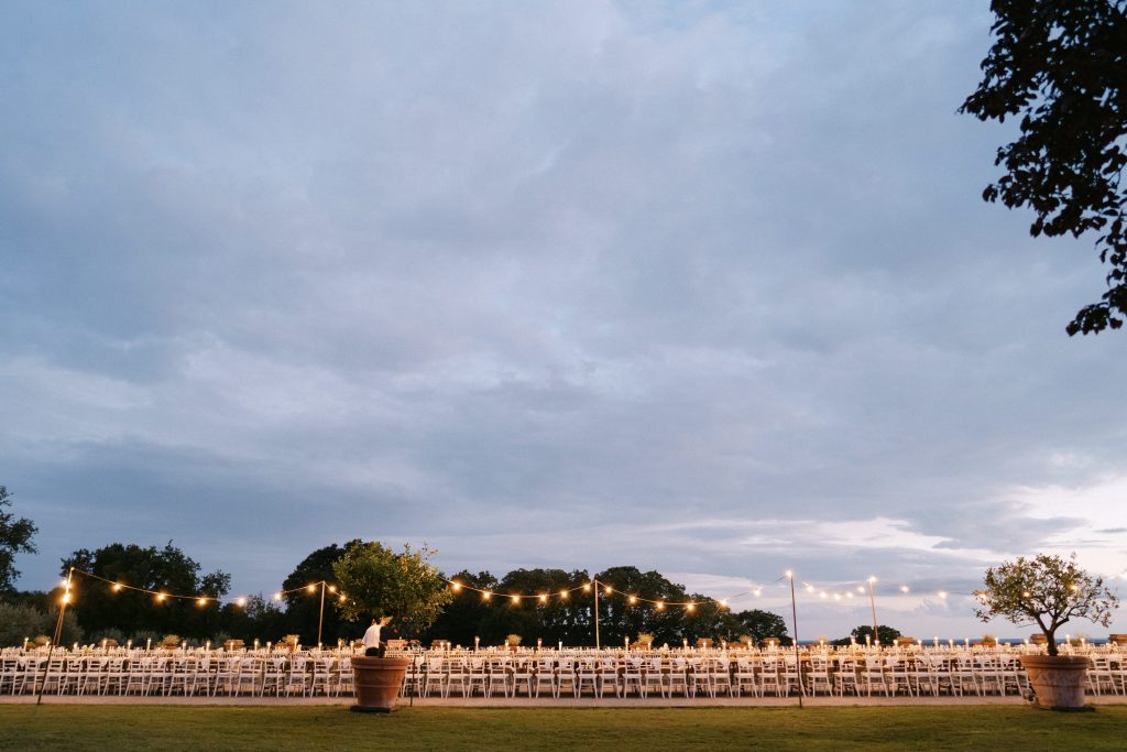 In questa foto, parte della serie matrimoni 2025 di Ilaria & Andrea, la lunga tavolata del ricevimento nuziale è allestita all’aperto sotto un cielo al tramonto. File ordinate di sedie bianche e tavoli decorati con luci calde e candele creano un’atmosfera elegante e intima. Il grande prato, gli alberi in vaso e le luci sospese tra i pali evocano un incanto rustico e raffinato.