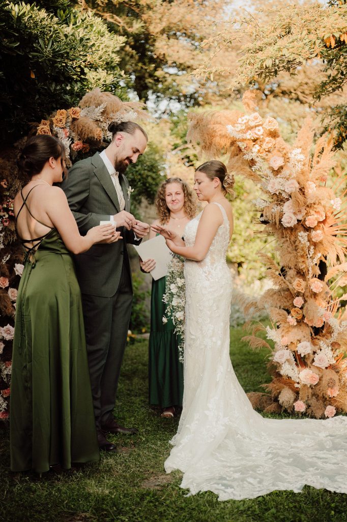 In questa foto, gli sposi si scambiano le fedi durante la cerimonia all’aperto, circondati da testimoni e amici. Il momento è intimo e carico di emozione, con un arco floreale alle loro spalle decorato con pampas, rose e fiori nei toni caldi. Un dettaglio poetico dei matrimoni 2025 di Ilaria & Andrea.