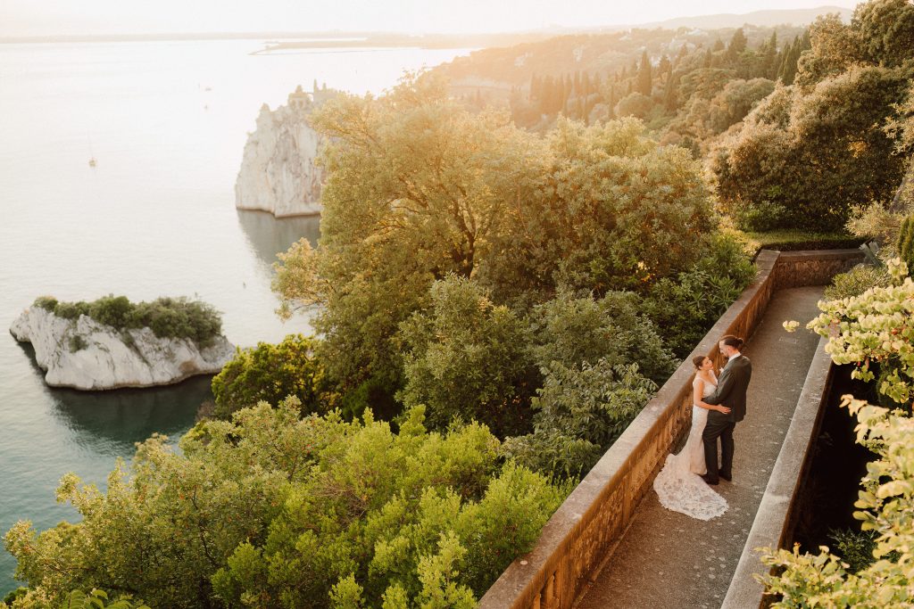 In questa foto, gli sposi si abbracciano su una terrazza panoramica immersa nel verde, con vista mozzafiato sul mare e su una scogliera. Il tramonto avvolge la scena in una luce dorata, regalando uno scatto romantico che racconta l’essenza dei matrimoni 2025 di Ilaria & Andrea.
