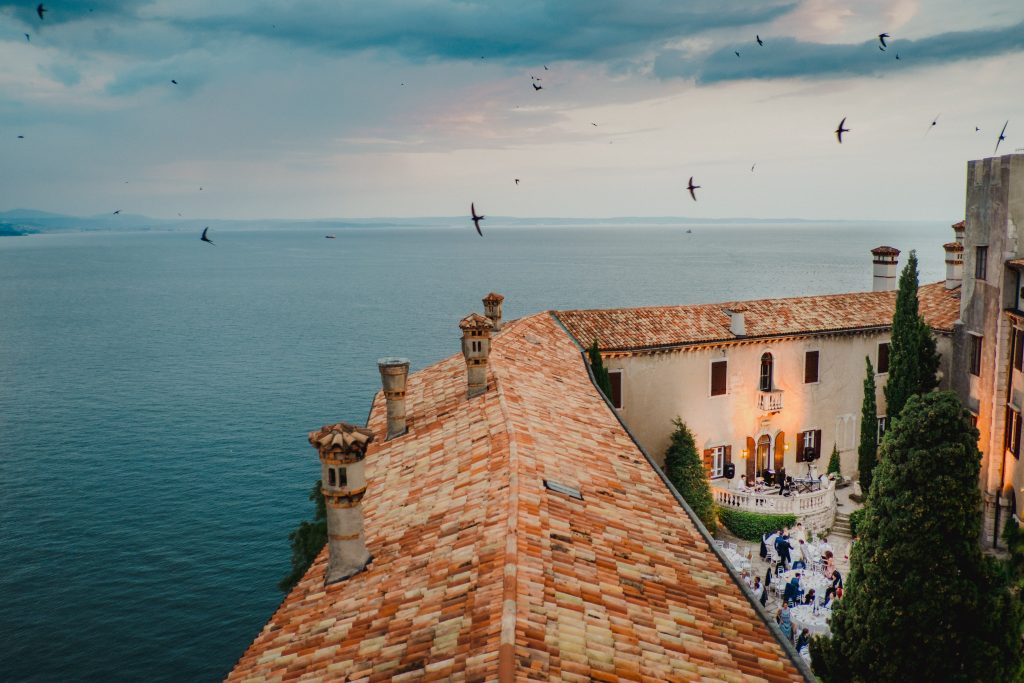 In questa foto, si vede una vista aerea della location del ricevimento con tetti in terracotta e il mare sullo sfondo. In basso, il cortile ospita i tavoli e gli invitati alla festa, illuminati da luci calde. Un’atmosfera suggestiva che racconta le atmosfere dei matrimoni 2025 di Ilaria & Andrea.