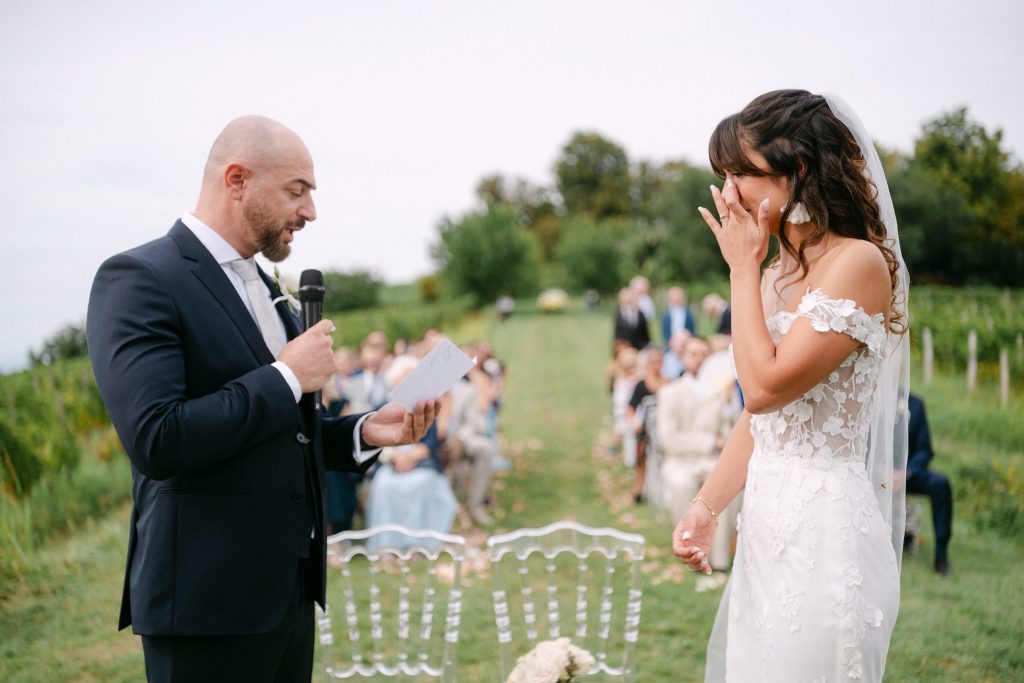 In questa foto, scattata durante i matrimoni 2025 di Ilaria & Andrea, lo sposo legge le sue promesse tenendo un microfono in mano, mentre la sposa, profondamente commossa, si asciuga le lacrime. Indossa un abito con spalle scoperte e dettagli floreali in pizzo. Sullo sfondo si intravedono gli ospiti seduti all’aperto, immersi nel verde.