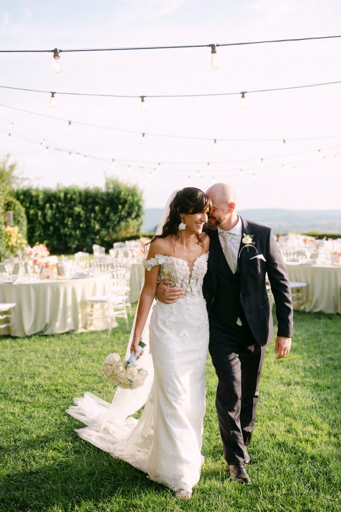 In questa foto dei matrimoni 2025 di Ilaria & Andrea, gli sposi camminano sorridenti, abbracciati, in un prato decorato con luci sospese. La sposa indossa un elegante abito bianco a sirena con ricami floreali e porta in mano un bouquet di rose chiare. L’atmosfera è romantica e rilassata, con i tavoli del ricevimento allestiti sullo sfondo.
