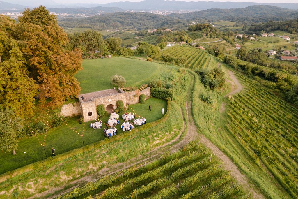 In questa foto dei matrimoni 2025 di Ilaria & Andrea, una vista aerea mostra la location immersa nel verde: una terrazza naturale tra i filari, con tavoli apparecchiati e luci sospese, incorniciata da un paesaggio collinare mozzafiato. Il ricevimento si svolge in un contesto intimo e suggestivo, circondato da natura e silenzio.