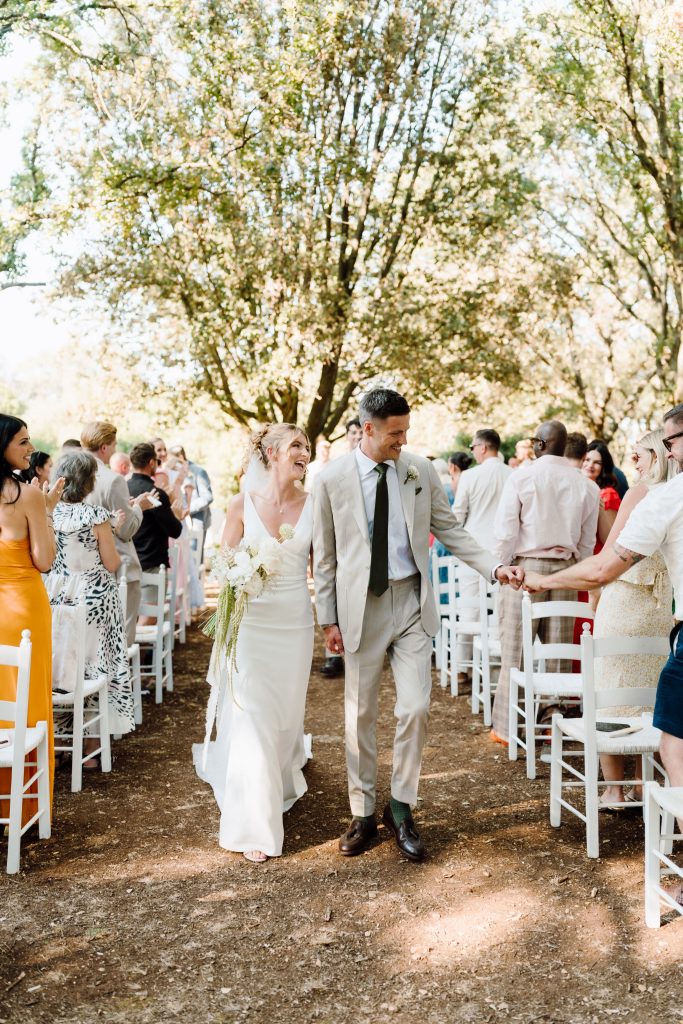 In questa foto dei matrimoni 2025 di Ilaria e Andrea, gli sposi camminano mano nella mano lungo il corridoio dell’allestimento nuziale all’aperto, tra due file di sedie bianche occupate dagli invitati che applaudono sorridenti. Ilaria indossa un abito bianco semplice e raffinato con scollo a V, mentre Andrea porta un completo chiaro con cravatta scura. Il sole filtra tra gli alberi, creando un’atmosfera calda e romantica, perfetta per i matrimoni 2025 di Ilaria & Andrea.