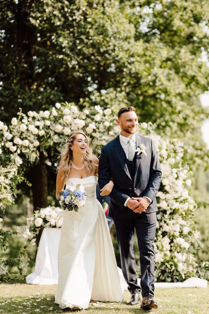 In questa foto, una coppia di sposi sorride e cammina all’aperto, appena dopo la cerimonia. Lei indossa un elegante abito bianco con scollo dritto e perle, mentre lui è in completo scuro con boutonnière bianca. Sullo sfondo si intravede un arco floreale bianco immerso nel verde. La scena fa parte del reportage matrimoni 2025 di Ilaria & Andrea.
