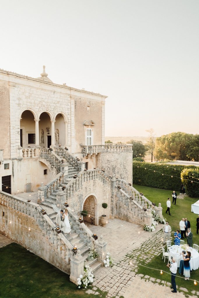 In questa foto, gli sposi scendono insieme una scenografica scalinata in pietra, circondata da decorazioni floreali bianche e luci sospese. Sullo sfondo si staglia una maestosa villa antica con arcate, tipica dell’Italia meridionale. L’immagine cattura la bellezza senza tempo della location scelta per uno dei matrimoni 2025 di Ilaria & Andrea.