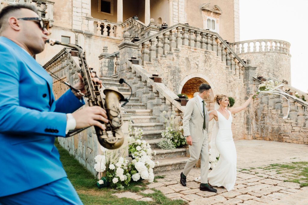 In questa foto, Ilaria e Andrea si godono un momento festoso all’esterno della villa storica, mentre un sassofonista in abito blu elettrico suona in primo piano. Ilaria alza il bouquet con gioia, sorridendo al pubblico, mentre Andrea la osserva con affetto. L’atmosfera è vivace, elegante e piena di energia, proprio come nei matrimoni 2025 di Ilaria & Andrea.