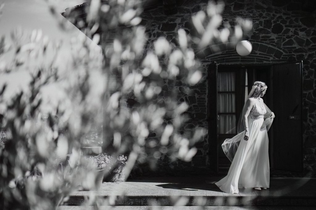 In questa foto, scattata durante i matrimoni 2025 di Ilaria & Andrea, la sposa è ritratta da sola in un’elegante immagine in bianco e nero. Attraverso i rami sfocati di un ulivo, si intravede Ilaria che, con il velo sollevato dal vento, cammina con grazia davanti a una struttura in pietra, come uscita da un sogno toscano.