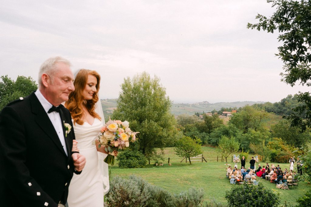 In questa foto, la sposa dei matrimoni 2025 di Ilaria & Andrea cammina emozionata a braccetto con un uomo più anziano, probabilmente il padre. Indossa un abito bianco con spalle scoperte, scollo a cuore e maniche scese. Il tessuto è morbido e avvolgente, con una linea dritta che valorizza la figura. Tiene in mano un bouquet dai toni pastello con rose gialle e fiori delicati. Sullo sfondo si intravede un paesaggio verde e collinare.