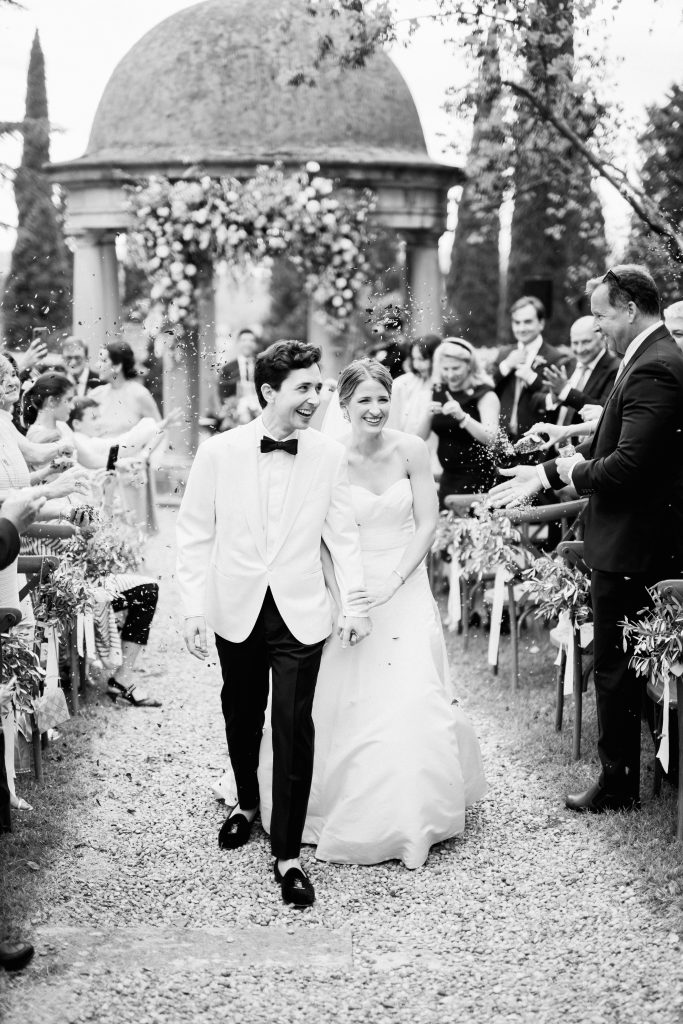 In questa foto in bianco e nero, parte della serie dedicata ai matrimoni 2025 di Ilaria & Andrea, la coppia appena sposata cammina felice lungo il corridoio all’aperto tra due file di ospiti sorridenti che lanciano petali o coriandoli. La sposa indossa un abito bianco senza spalline, con corpetto strutturato e gonna ampia, mentre lo sposo è elegante in smoking chiaro con papillon nero. La scena è gioiosa e immersa nel verde, con un romantico tempietto fiorito sullo sfondo.