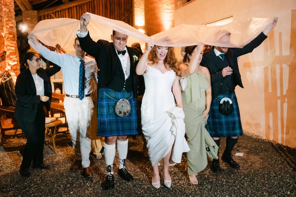 In questa foto, durante i festeggiamenti dei matrimoni 2025 di Ilaria & Andrea, la sposa e lo sposo ridono mentre partecipano a un gioco tradizionale, sollevando insieme un telo sopra le loro teste. L’abito da sposa mostra un dettaglio inedito: la parte frontale è sollevata leggermente, rivelando delle file di bottoncini e scarpe bianche a punta. La scena è vivace, con amici e parenti che partecipano al momento con entusiasmo.