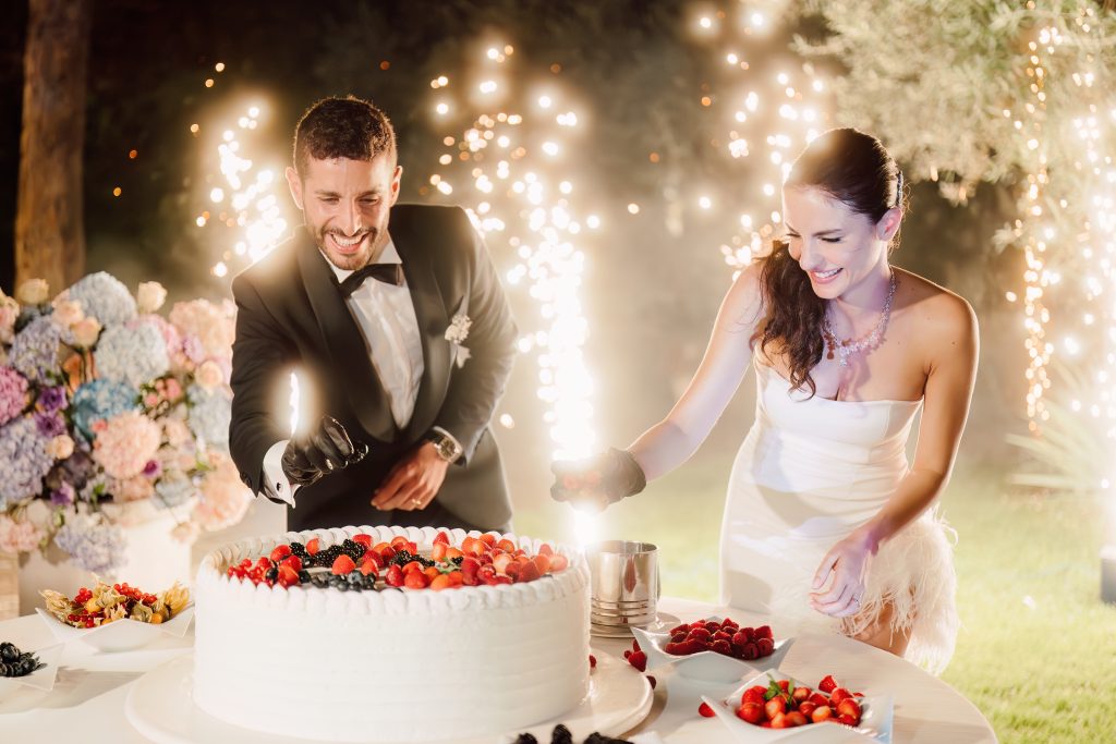 In questa foto, la coppia di sposi sorride mentre accende insieme due scintille davanti alla torta nuziale durante il taglio torta serale. Lei indossa un abito corto con piume, lui uno smoking elegante. Sullo sfondo, esplosioni luminose e fiori colorati creano un’atmosfera di festa. Uno scatto gioioso dai matrimoni 2025 di Ilaria & Andrea.