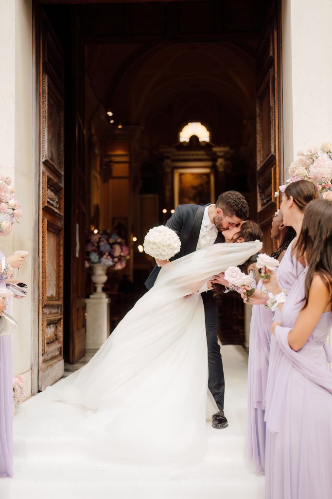 In questa foto, gli sposi si scambiano un bacio appassionato all’uscita della chiesa, circondati dalle damigelle vestite di lilla. La sposa, con un abito vaporoso e un bouquet di rose bianche, viene dolcemente piegata all’indietro dallo sposo in un gesto romantico. L’immagine, tratta dai matrimoni 2025 di Ilaria & Andrea, cattura l’emozione del momento.