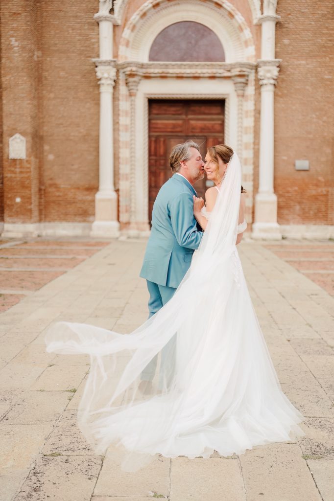 In questa foto, gli sposi si abbracciano sorridenti davanti al portone di una chiesa antica. L’abito della sposa si muove con il vento, creando un effetto etereo. Lo sposo indossa un completo azzurro polvere. La scena trasmette complicità e dolcezza, tipica dello stile intimo ed elegante dei matrimoni 2025 di Ilaria & Andrea.