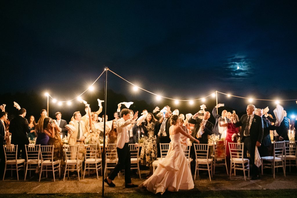 In questa foto dei matrimoni 2025 di Ilaria & Andrea, la scena si svolge di notte sotto una fila di luci sospese. Gli ospiti, in piedi e sorridenti, sventolano tovaglioli in aria mentre la coppia di sposi balla al centro, accolta con entusiasmo. La sposa indossa un abito bianco con strascico lo sposo danza con disinvoltura. L’atmosfera è vibrante, festosa, piena di vita.