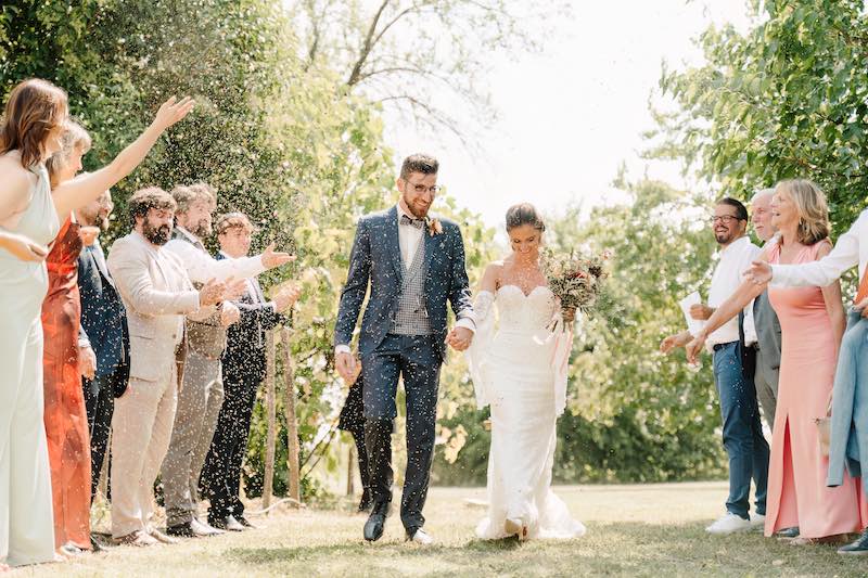 In questa foto, la coppia di sposi cammina mano nella mano mentre gli invitati, disposti in due file, lanciano petali o riso festosamente. Lo sfondo verde e il sole creano un’atmosfera gioiosa e spontanea. Una scena vibrante tipica dei matrimoni 2025 di Ilaria & Andrea.