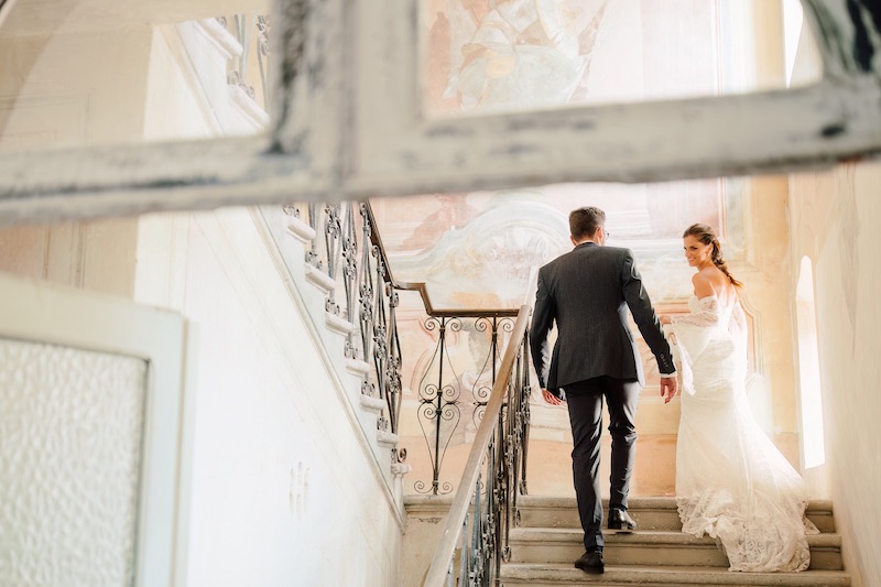 In questa foto, una coppia di sposi eleganti sale una scalinata antica circondata da affreschi e luce naturale. La sposa, in un abito bianco con maniche in pizzo, guarda sorridendo verso la fotocamera. L’atmosfera è intima e luminosa. Uno scatto raffinato che racconta l’eleganza dei matrimoni 2025 di Ilaria & Andrea.