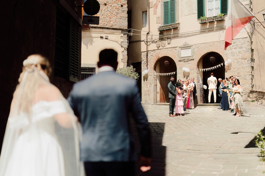 In questa foto, parte del racconto visivo dei matrimoni 2025 di Ilaria & Andrea, la sposa avanza verso il luogo della cerimonia, accompagnata da una figura maschile di spalle. Davanti a loro, amici e familiari li attendono con sorrisi e fiori, all’ingresso di una chiesa dal sapore antico, decorata con festoni bianchi. La scena è carica di attesa e commozione.
