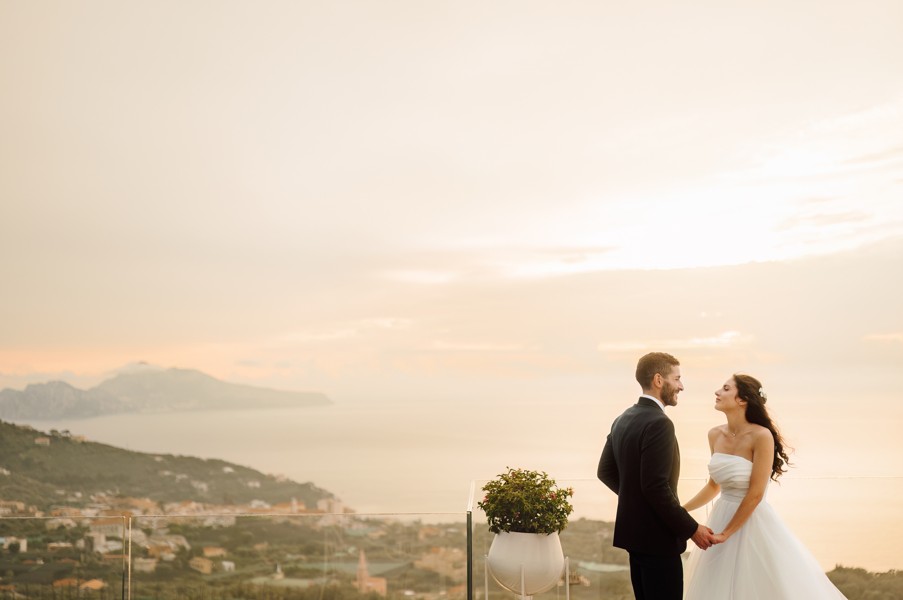 In questa foto, una coppia di sposi si tiene per mano su una terrazza panoramica al tramonto, con lo sguardo perso l’uno nell’altra. Lo sfondo è una distesa dorata che si perde all’orizzonte, mentre il cielo sfuma tra rosa e arancio. La scena, parte dei matrimoni 2025 di Ilaria & Andrea, trasmette un’intima serenità.