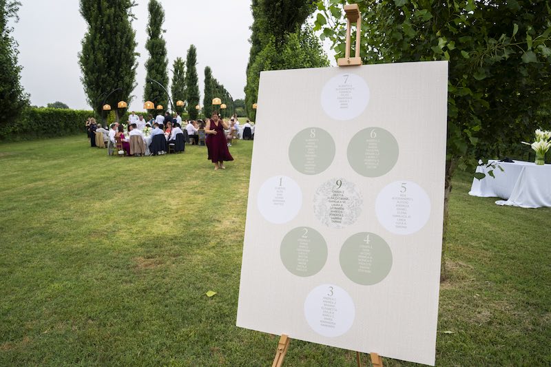 In questa foto, in primo piano un tableau de mariage  che mostra la disposizione dei tavoli rotondi a rombo e sullo sfondo gli invitati di un matrimonio seduti ai tavoli