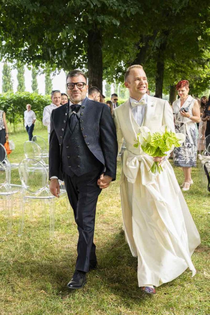 In questa foto, l'uscita dal luogo della cerimonia di due sposi. Uno indossa un abito da sposa damascato con giacca dallo scollo coreano; l'altro indossa un abito da sposo di colore bianco con giacca e ampia gonna