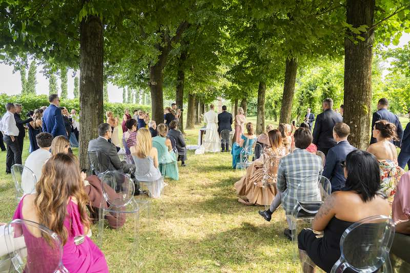 In questa foto, un momento della cerimonia del matrimonio di Alberto e Aldis progettato da OttoQuattro Eventi su un prato sotto ad un filare d'alberi