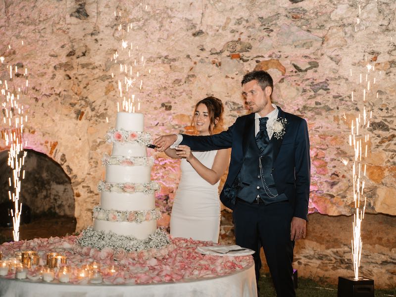 In questa foto, due sposi tagliano una torta nuziale a piani di colore bianco, decorata con fiori. Sul tavolo sono presenti petali di colore rosa e candele di light. Alle loro spalle esplodono fuochi d'artificio