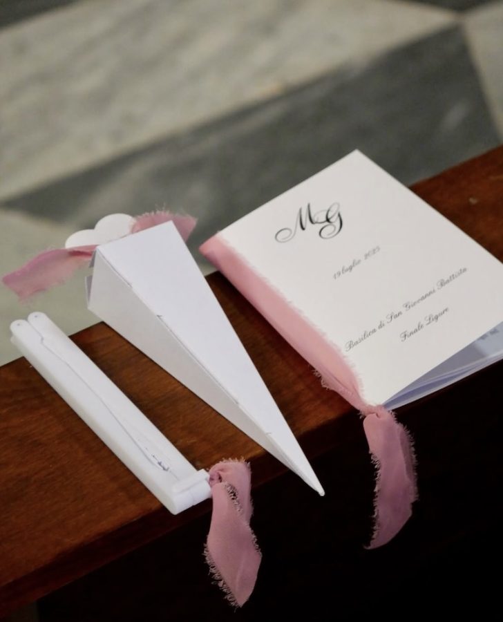In questa foto, il dettaglio della stationery ideata da Valeria Fazio composta da: un ventaglio, un cono portariso e un libretto messa di colore bianco con nastri di garza di colore rosa peonia
