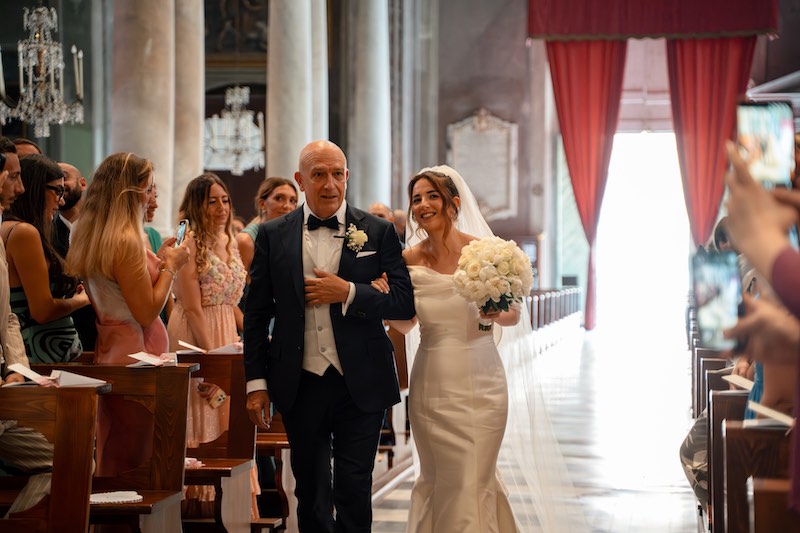 In questa foto, una sposa entra in chiesa al braccio del papà. La sposa stringe tra le mani un bouquet Total White di rose e peonie