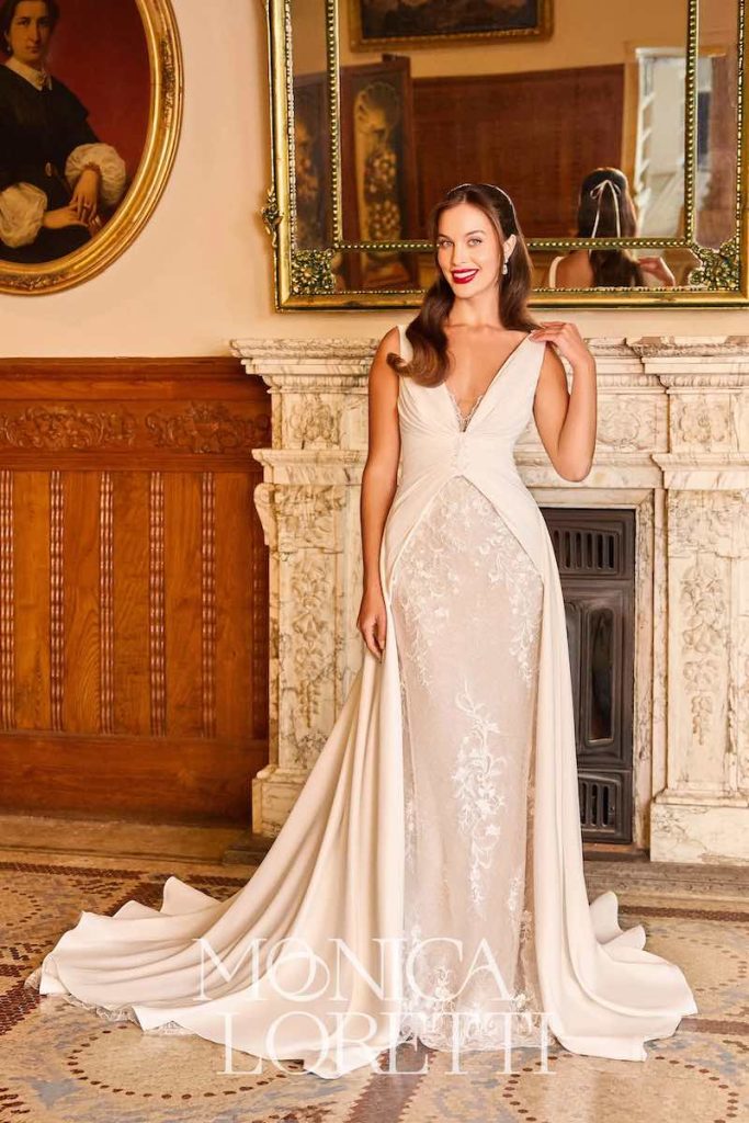 In questa foto, una modella posa indossando un abito da sposa Monica Loretti dalla silhouette a semi sirena in pizzo con giacca lunga fino a diventare una lunga coda in cady