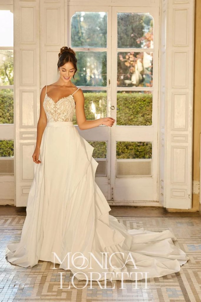 In questa foto, una modella posa indossando un abito da sposa Monica Loretti 2026 con silhouette scivolata con gonna in cady e corpetto in pizzo con spalline sottili
