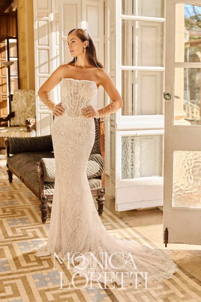 In questa foto, una modella posa indossando un abito da sposa Monica Loretti con silhouette a semi sirena in pizzo con ricami e guanti abbinati 