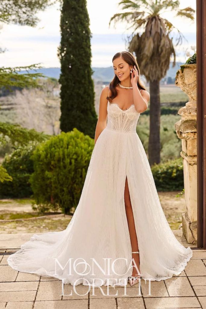 In questa foto, una modella posa indossando un abito da sposa Monica Loretti 2026 dalla silhouette ad A con gonna in pizzo, spacco frontale e corpetto steccato con scollo a cuore