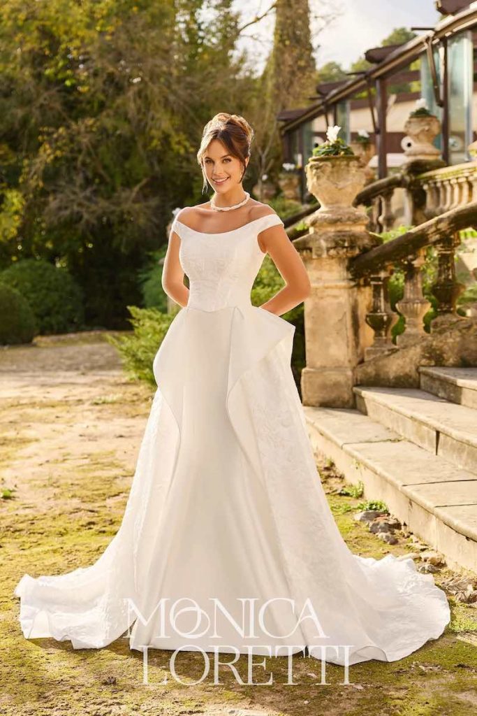 In questa foto, una modella posa indossando un abito da sposa Monica Loretti 2026 con scollo omerale e sovragonna in tessuto jacquard