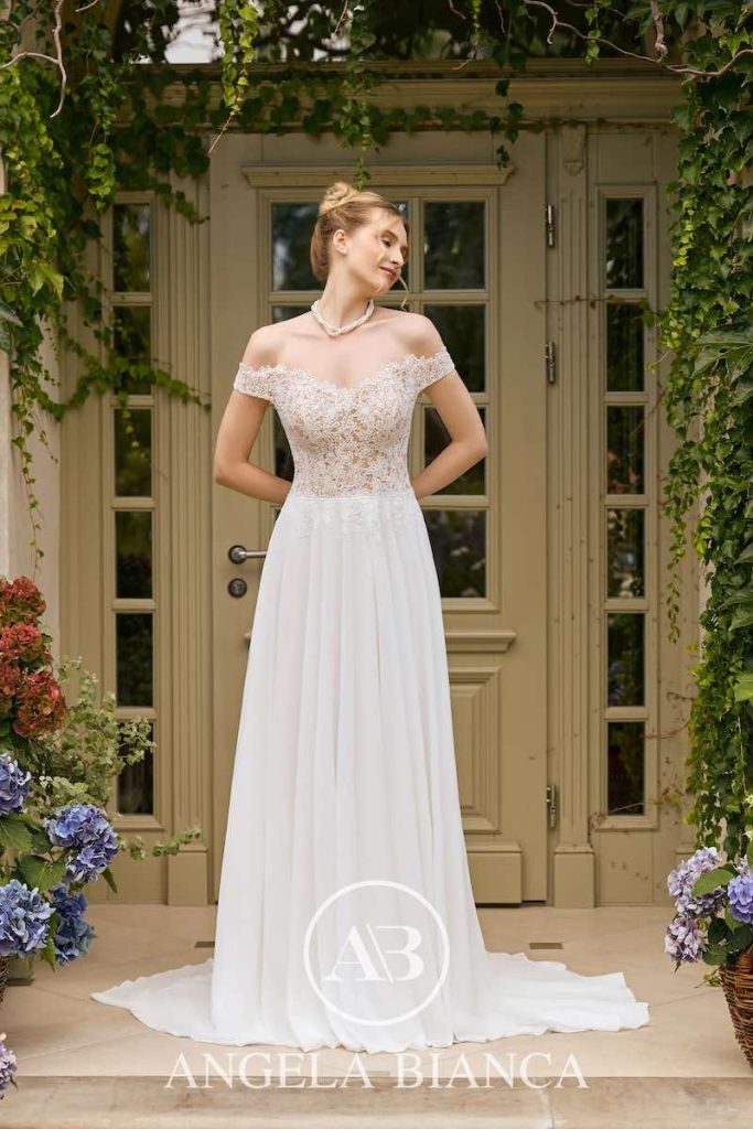 In questa foto, una modella posa indossando un abito da sposa Angela Bianca 2026 con silhouette scivolata in cady e corpetto in pizzo floreale