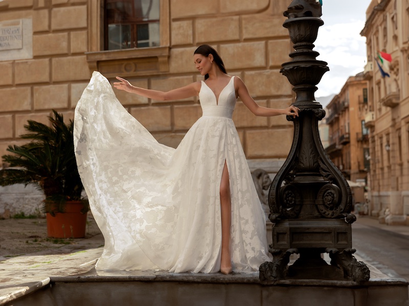 In questa foto, una modella posa indossando un abito da sposa Monica Loretti 2026 dalla silhouette ad A, spacco, intarsi floreali e ampio scollo a V