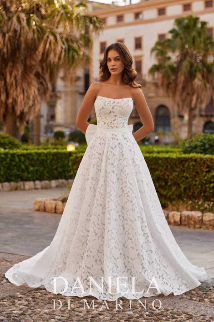 In questa foto, una modella posa indossando un abito da sposa Daniela Di Marino 2026 con silhouette con ricami floreali, scollatura dritta e fiocco posteriore