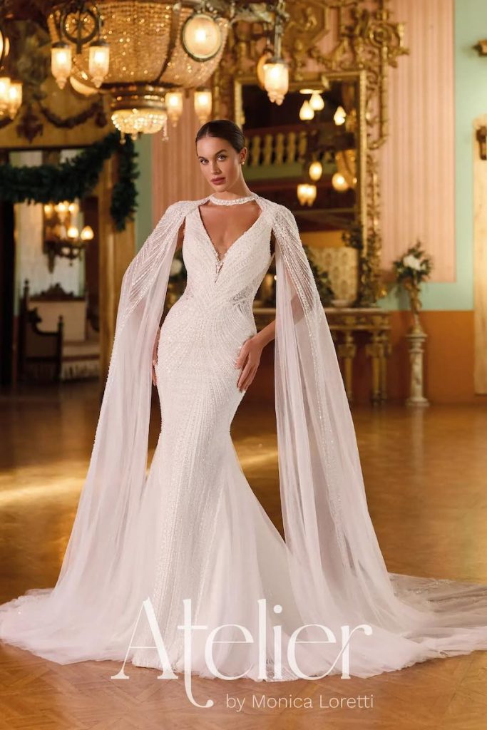 In questa foto, una modella posa indossando un abito da sposa Atelier by Monica Loretti con silhouette semisirena, ricami di perline e paillettes all over, scollo a V e cappa di tulle con ricami ramage