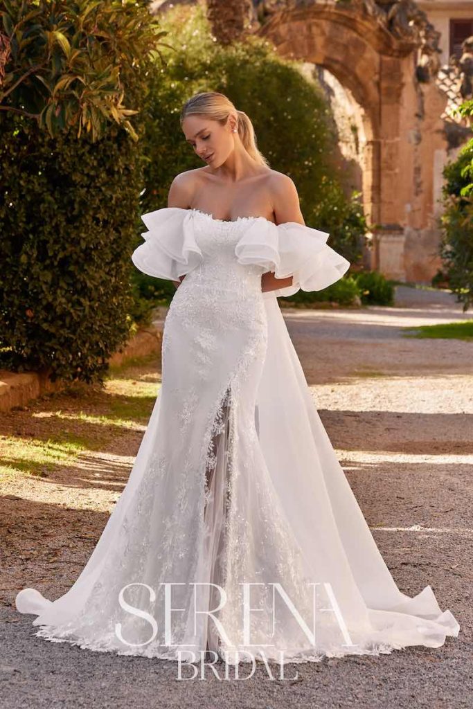 In questa foto, una modella posa indossando un abito da sposa Serena Bridal 2026 con silhouette a semi sirena, spacco frontale, scollo omerale e lungo strascico in organza