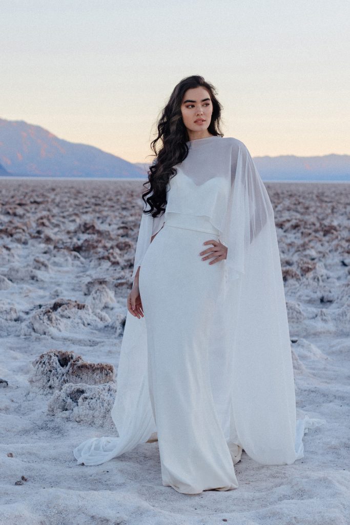 In questa foto si vede un abito da sposa con silhouette a colonna, realizzato da Allure Bridal in tessuto fluido e opaco che rappresenta una delle tendenze sposa 2026 . Il modello è caratterizzato da un’originale mantella in chiffon trasparente che avvolge le spalle e scende lungo i fianchi, creando un effetto etereo. Un look perfetto per chi sogna un look dallo stile moderno e minimale.