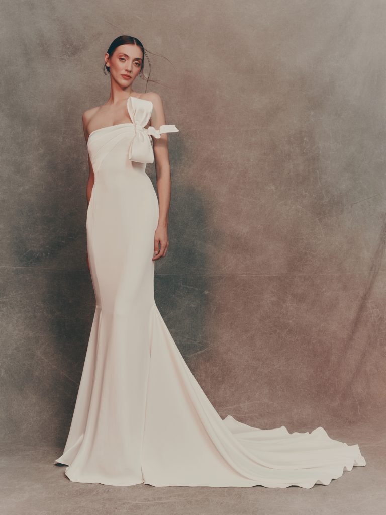 In questa foto si vede un abito da sposa Atelier Pronovias, dalla linea a sirena e taglio monospalla che rappresenta una delle tendenze sposa 2026. Realizzato in tessuto liscio, il vestito è impreziosito da un grande fiocco asimmetrico applicato sulla spalla, che dona un tocco couture al design essenziale. Lo strascico morbido completa l’eleganza pulita dell’insieme.