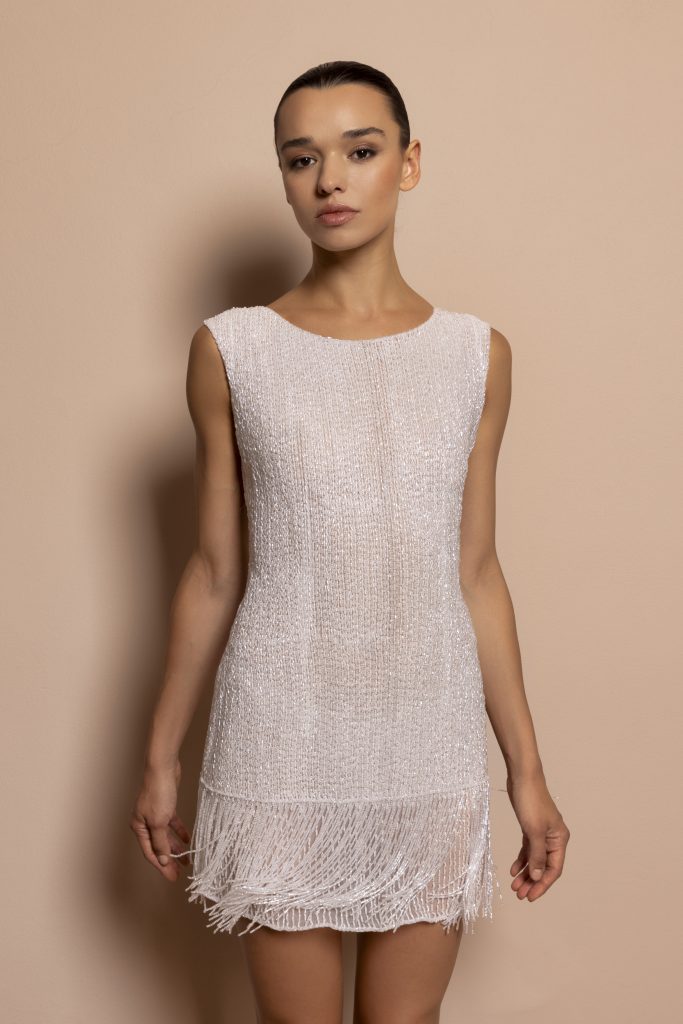 In questa foto si vede un abito da sposa corto della designer Daniela De Souza, dal design essenziale e sofisticato che rappresenta una delle tendenze sposa 2026 . Il vestito è interamente ricoperto di micro-perline luminose, con una lavorazione verticale che slancia la figura. Il fondo è decorato con frange leggere che donano movimento e un tocco rétro contemporaneo.