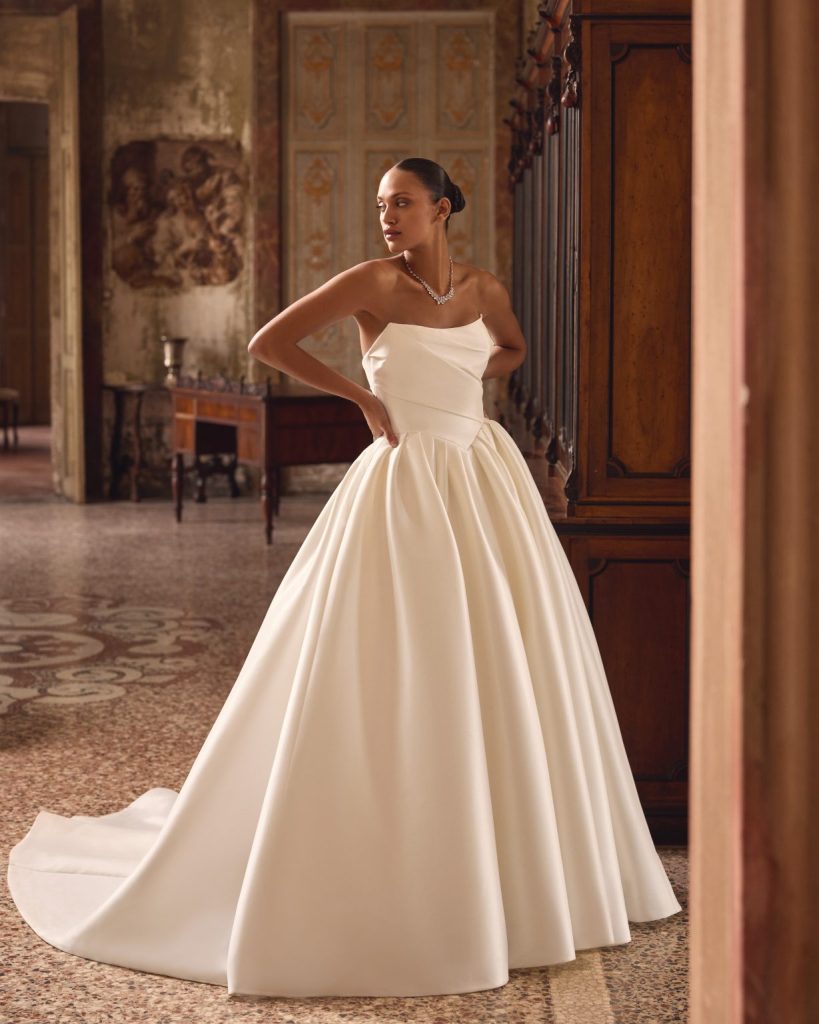 In questa foto, la sposa indossa un abito da sposa principesco in seta che rappresenta una delle tendenze sposa 2026 , caratterizzato da un corpetto strutturato senza spalline e da una scollatura dritta. Il punto vita è messo in risalto da un taglio geometrico che si apre in una gonna ampia e voluminosa con morbide pieghe, creando un effetto regale ed elegante. Lo strascico lungo completa il look con maestosità.