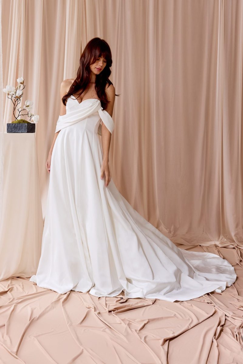 In questa foto si vede un abito realizzato da Nagae dal design elegante e scultoreo, con corpetto drappeggiato che incrocia sul busto e si trasforma in maniche morbide. La gonna ampia e fluida scende in una cascata di tessuto, creando uno stile sofisticato e poetico. Il tessuto lucido e la linea pulita conferiscono una raffinatezza senza tempo.