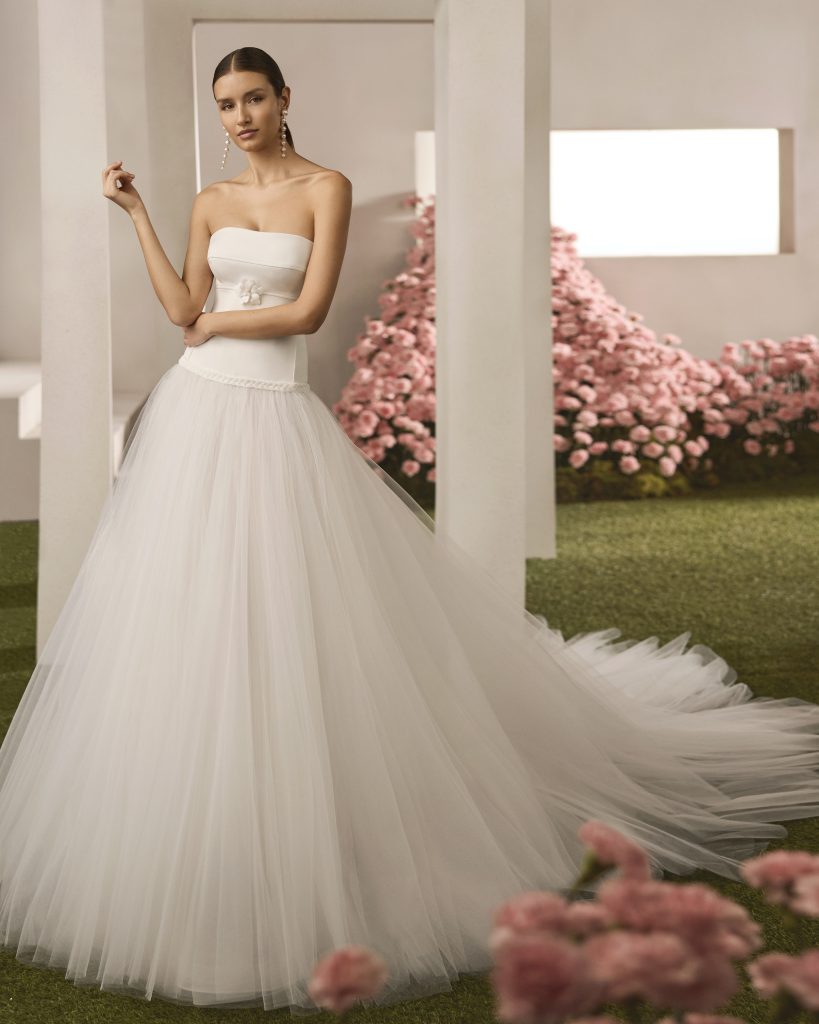 In questa foto si vede un abito da sposa che rappresenta una delle tendenze sposa 2026 firmato Rosa Clarà Couture con bustino liscio e minimal, unito a una gonna estremamente ampia e fluida in tulle bianco. La linea a vita basca e l'applicazione floreale tono su tono che sottolinea il punto vita con discrezione. Il look è impreziosito da orecchini pendenti lunghi.